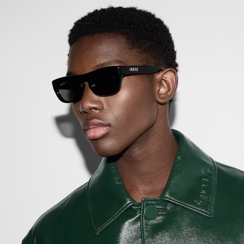 Close-up of person wearing black Gucci sunglasses and a dark green leather jacket against white background